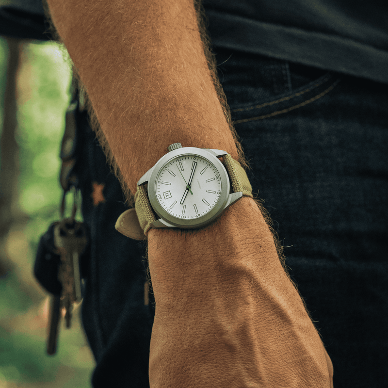 Big Idea Design LLC titanium field watch on a wrist with khaki canvas strap and white dial outdoors